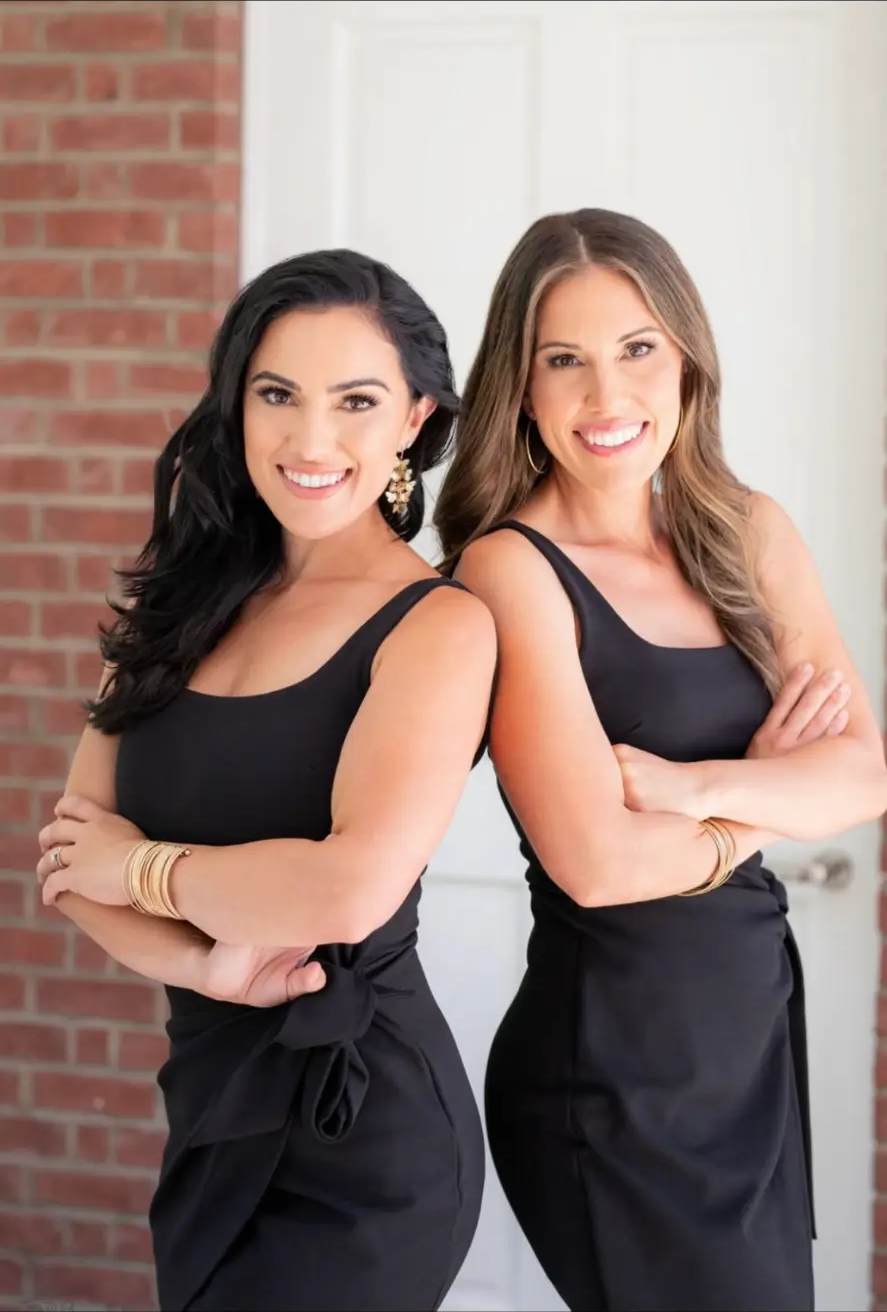 Julianna Sanchez and Cristina Nebot standing back-to-back in elegant black dresses with arms crossed, smiling